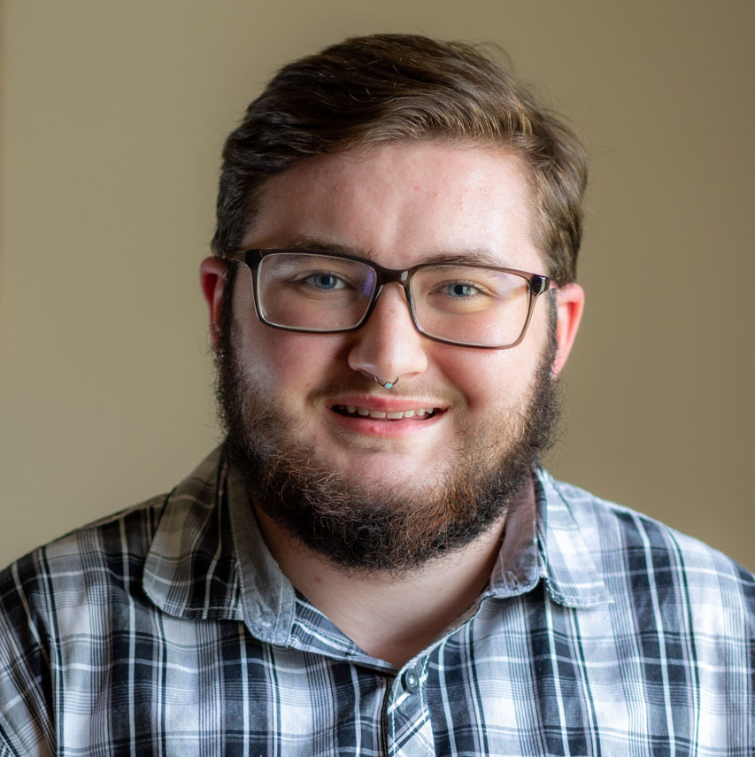 The image shows Charli, a white man with cropped brown hair and a short beard. He is wearing a gingham navy button-down shirt. He is also wearing rectangular brown glasses and has a nose piercing. He is smiling and standing against a tan background.