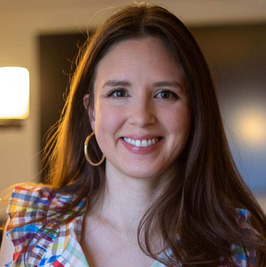 The image shows Lindsey, a white woman with long, straight, brown hair parted down the middle. She is wearing a rainbow gingham shirt, pink lipstick, and golden hoop earrings. She is smiling.